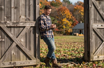 Farmer smiling at his phone while leaning against a barn door in warm afternoon light on a New England farm