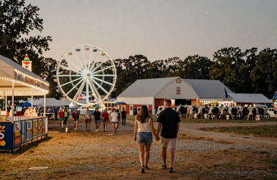 Couple walking through a North Carolina county fair at twilight with string lights and a ferris wheel in the background