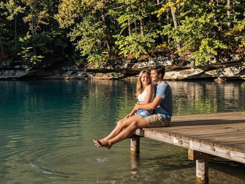 Happy couple sitting on a wooden dock beside a clear Ozark lake in Missouri at golden hour with limestone bluffs and forest