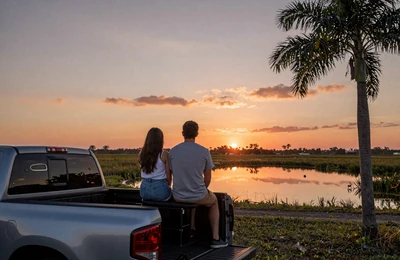 Couple watching sunset from a pickup truck tailgate over Florida farmland with palm tree silhouettes