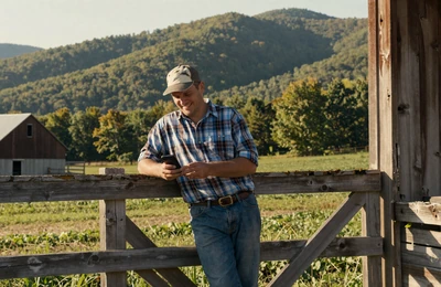 Farmer smiling at his phone while leaning against a barn door on a West Virginia mountain farm